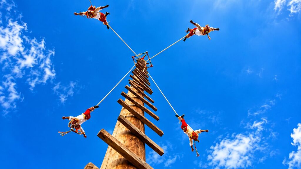 Spectacle traditionnel des Voladores de Papantla au Mexique, avec quatre danseurs en costumes colorés suspendus à des cordes autour d'un grand mât en bois.