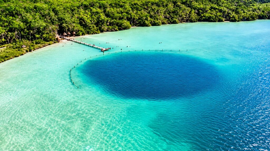 Vue aérienne d'un cénote aux eaux turquoise et bleu profond, entouré de végétation tropicale à Bacalar, au Mexique.