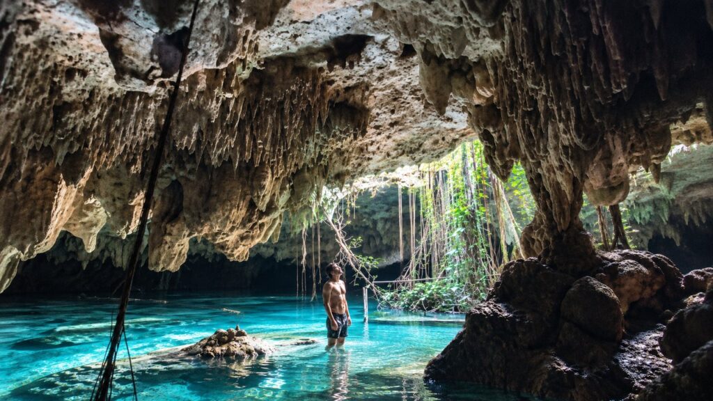 Homme debout dans les eaux turquoise d'un cénote souterrain du Yucatán, sous des stalactites et des racines tropicales.