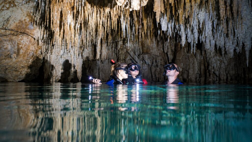Trois visiteurs explorent un cénote souterrain du Yucatán avec masques et lampes, sous de nombreuses stalactites calcaires se reflétant dans l'eau cristalline.