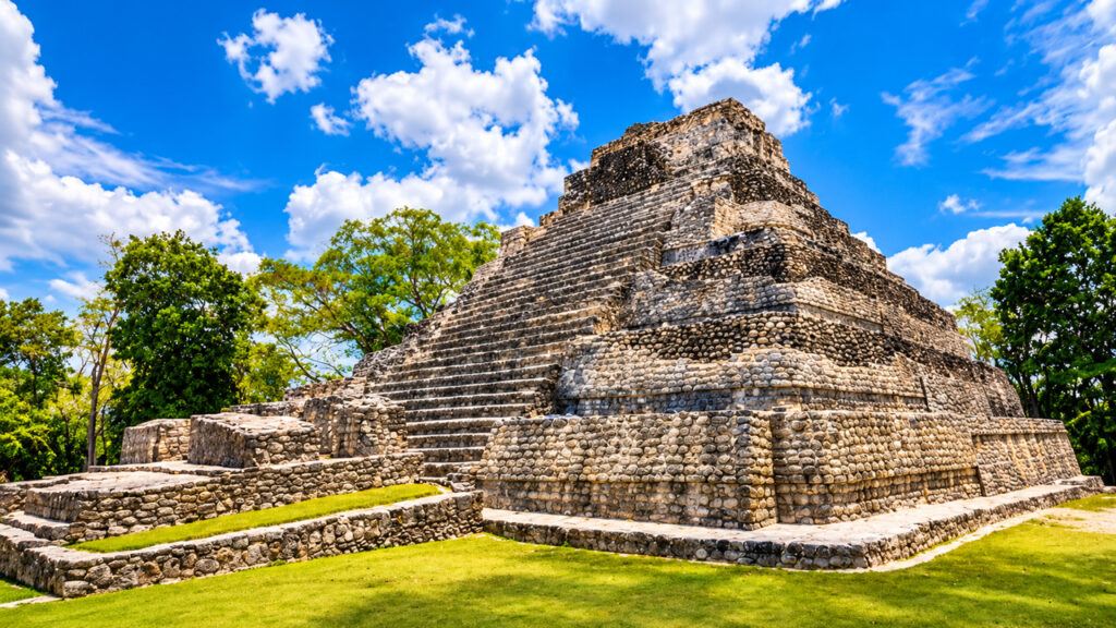 Pyramide maya en pierre avec un grand escalier central, entourée de végétation tropicale sous un ciel bleu au Mexique.